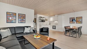 Una sala de estar moderna con sofá, mesa de centro y zona de comedor. Se observan armarios de cocina y una mesa de comedor con sillas.