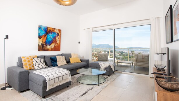 A modern living room with a gray sofa, a glass coffee table, and a large window offering a city view. - Blue Pearl 206 by Avecoeur (Nice)