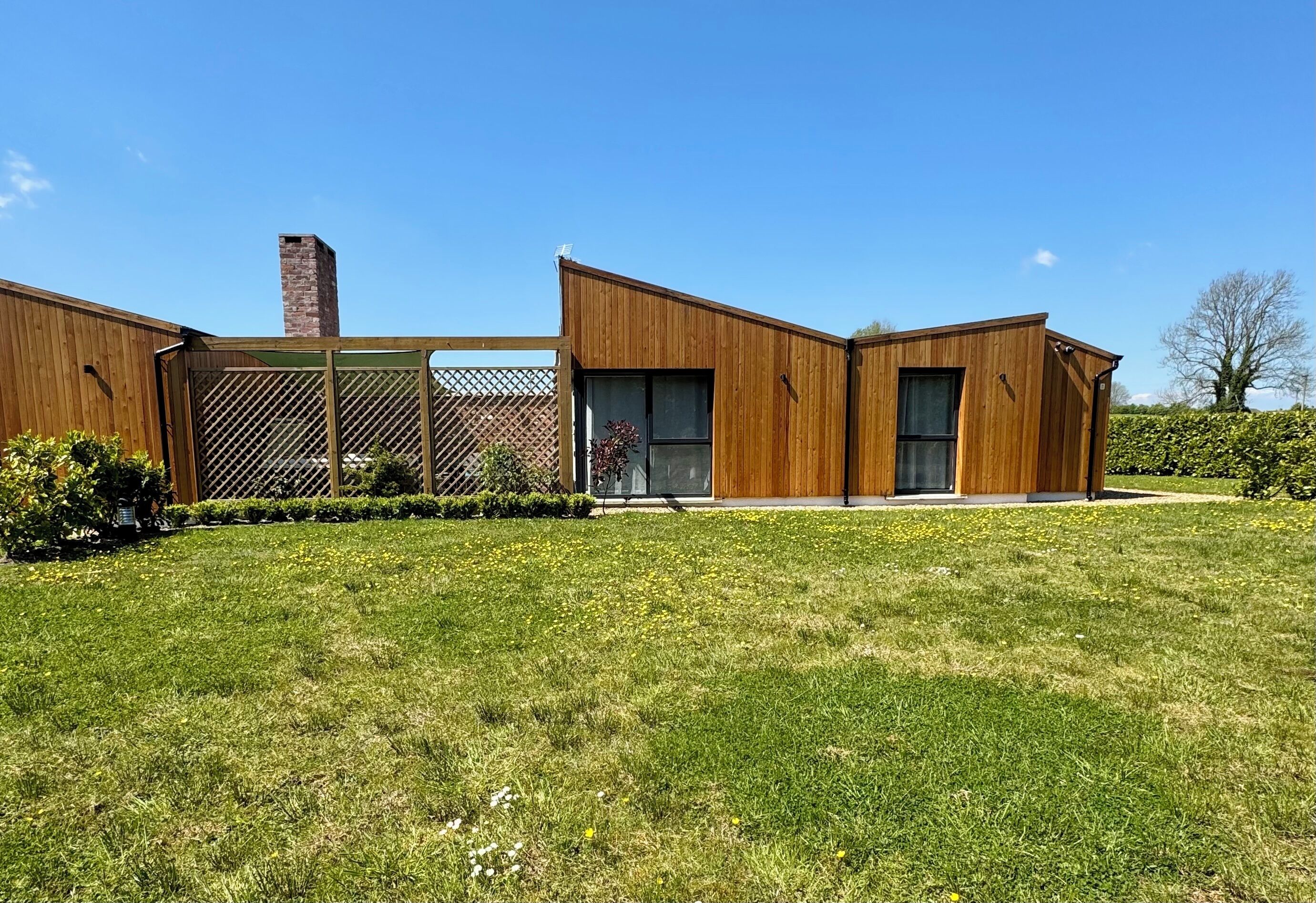 Una casa moderna de dos pisos con fachada de madera, una chimenea de ladrillo y un patio cercado con césped y plantas pequeñas.