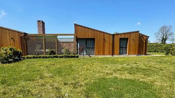 Una casa moderna de dos pisos con fachada de madera, una chimenea de ladrillo y un patio cercado con césped y plantas pequeñas.