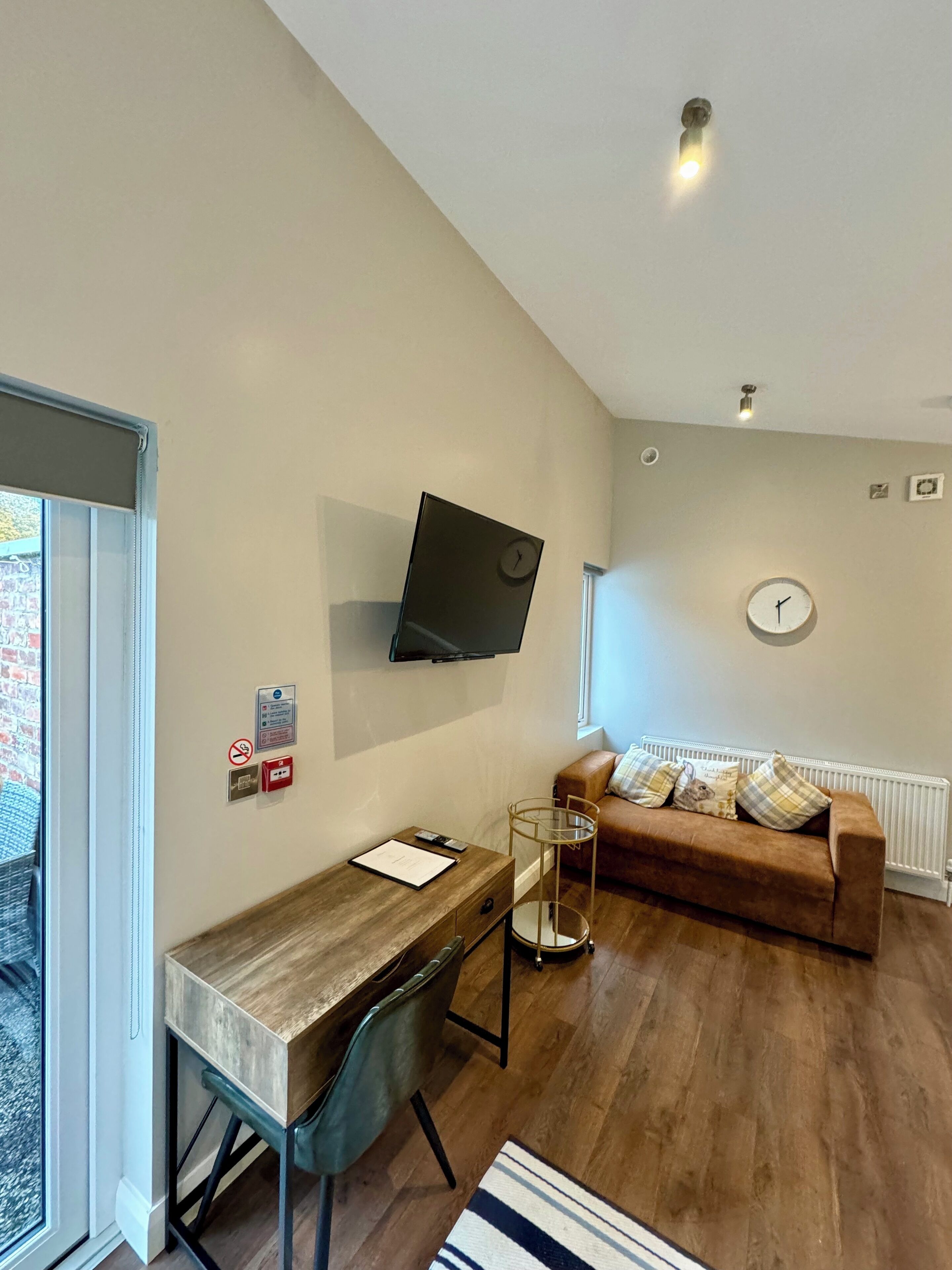 A modern living room with a wooden desk, a brown sofa, a mounted TV, and a window with a view of a brick building.