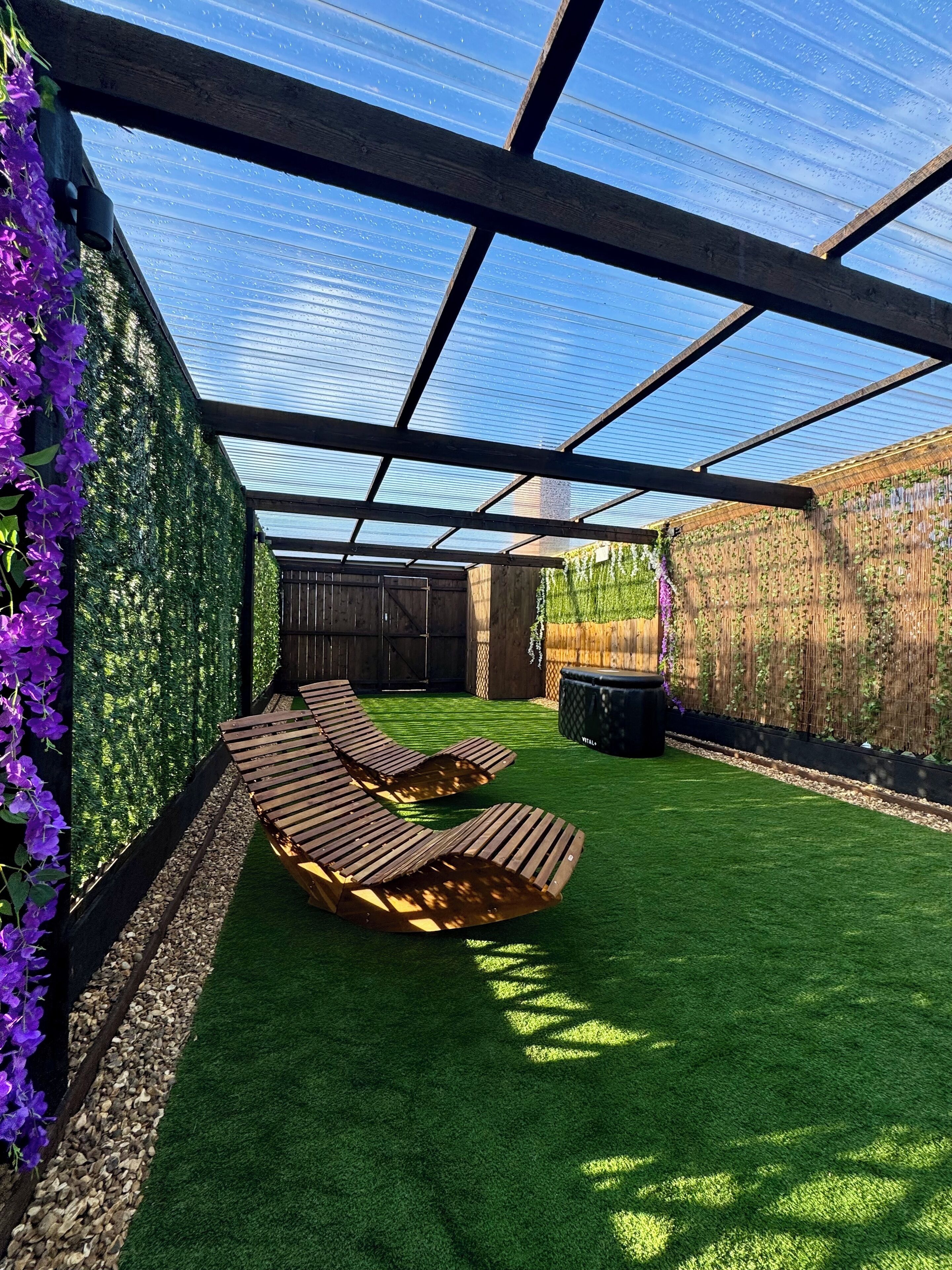A covered outdoor area with artificial grass, wooden loungers, and a pergola structure.