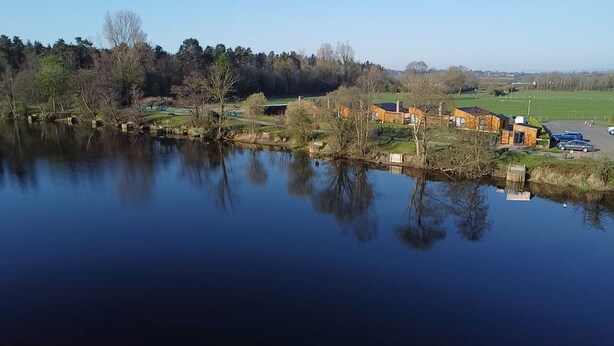 A calm lake with reflections of trees and buildings, surrounded by a park with a few structures and a parking area. - The River Bann Retreat (Ballymena)