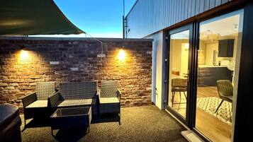 A patio area with wicker furniture and a brick wall, leading to a glass door entrance.