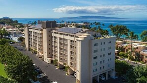 A multi-story hotel with a rooftop pool, surrounded by palm trees and a view of the ocean. - 180° Ocean views • Luxe condo • Steps to Beach (Kihei)