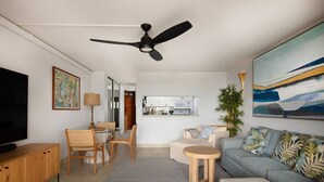 A modern living room with a ceiling fan, a television, a dining area with chairs, a sofa, and a kitchen in the background. - 180° Ocean views • Luxe condo • Steps to Beach (Kihei)