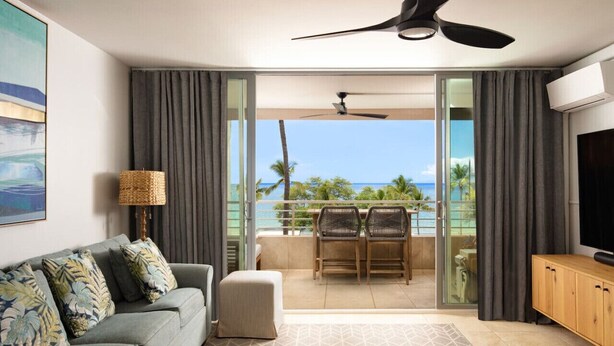 A modern living room with a sofa, a coffee table, and a TV. There is a balcony with a view of the ocean and palm trees. - 180° Ocean views • Luxe condo • Steps to Beach (Kihei)