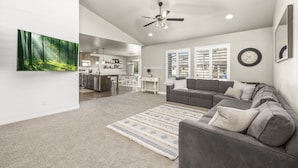 A modern living room with a gray sectional sofa, a large forest-themed painting, and a ceiling fan.