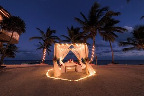 Escena de playa con una pérgola y luces colgantes, palmeras y vista al océano.