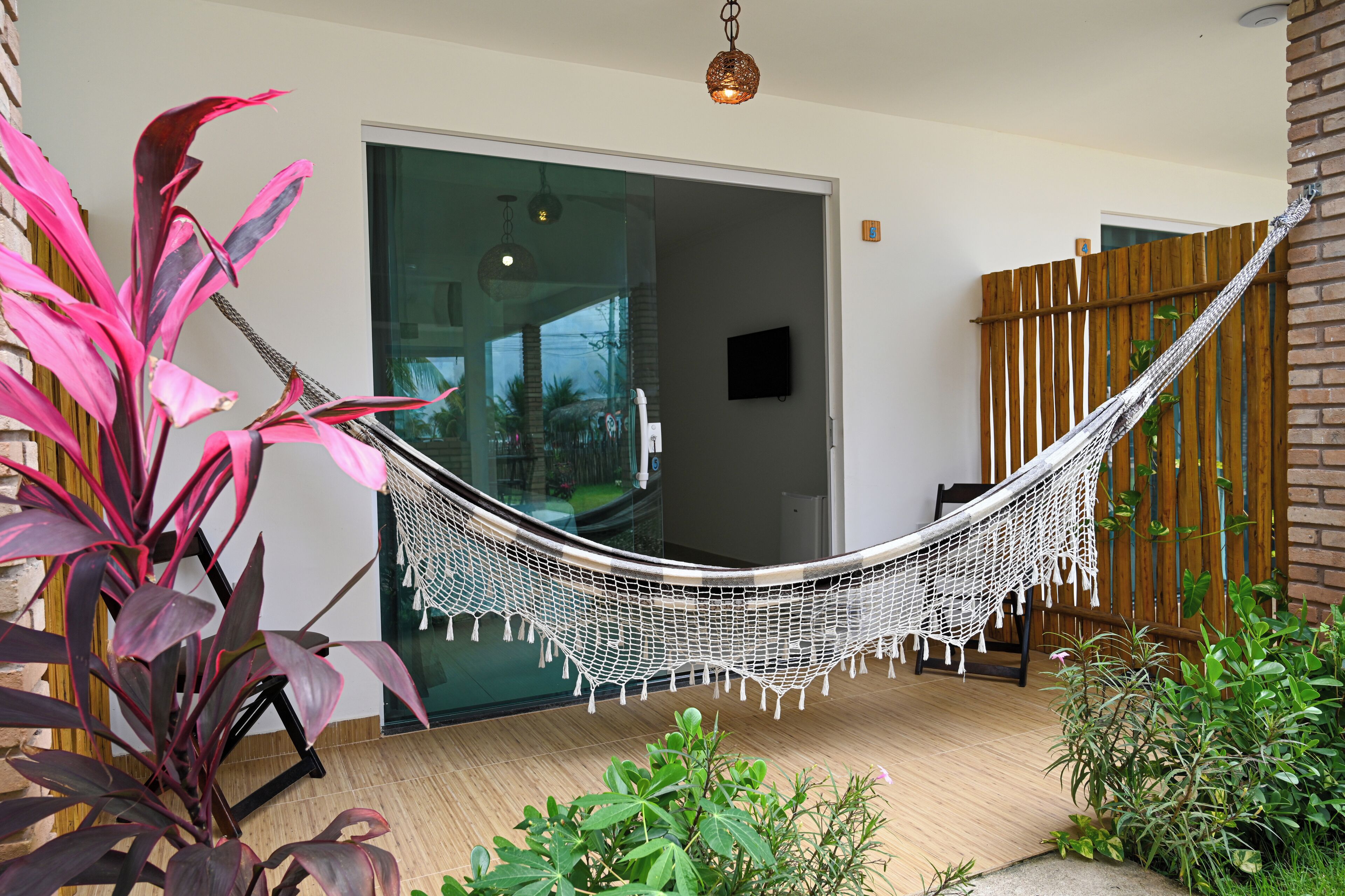 A hammock chair in a modern room with a glass door, a wooden fence, and various plants.