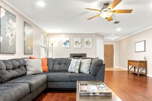 Un salon moderne avec un canapé d’angle, un plancher en bois, un ventilateur de plafond et des œuvres d’art accrochées aux murs.