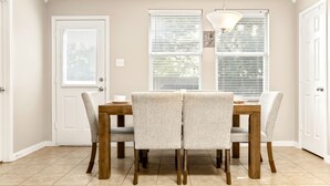 Un comedor con una mesa de madera y cuatro sillas, dos ventanas con persianas, y una lámpara colgante.