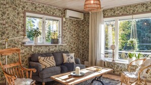 Ein gemütliches Wohnzimmer mit einem grauen Sofa, Holzstühlen, einem Holztisch und Blick ins Grüne durch große Fenster.