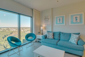A modern living room with a large window, a blue sofa, two blue chairs, a white coffee table, and framed artwork on the wall.