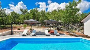 Une piscine entourée d’arbres et d’un mur de pierre, avec des chaises longues et des parasols à proximité.