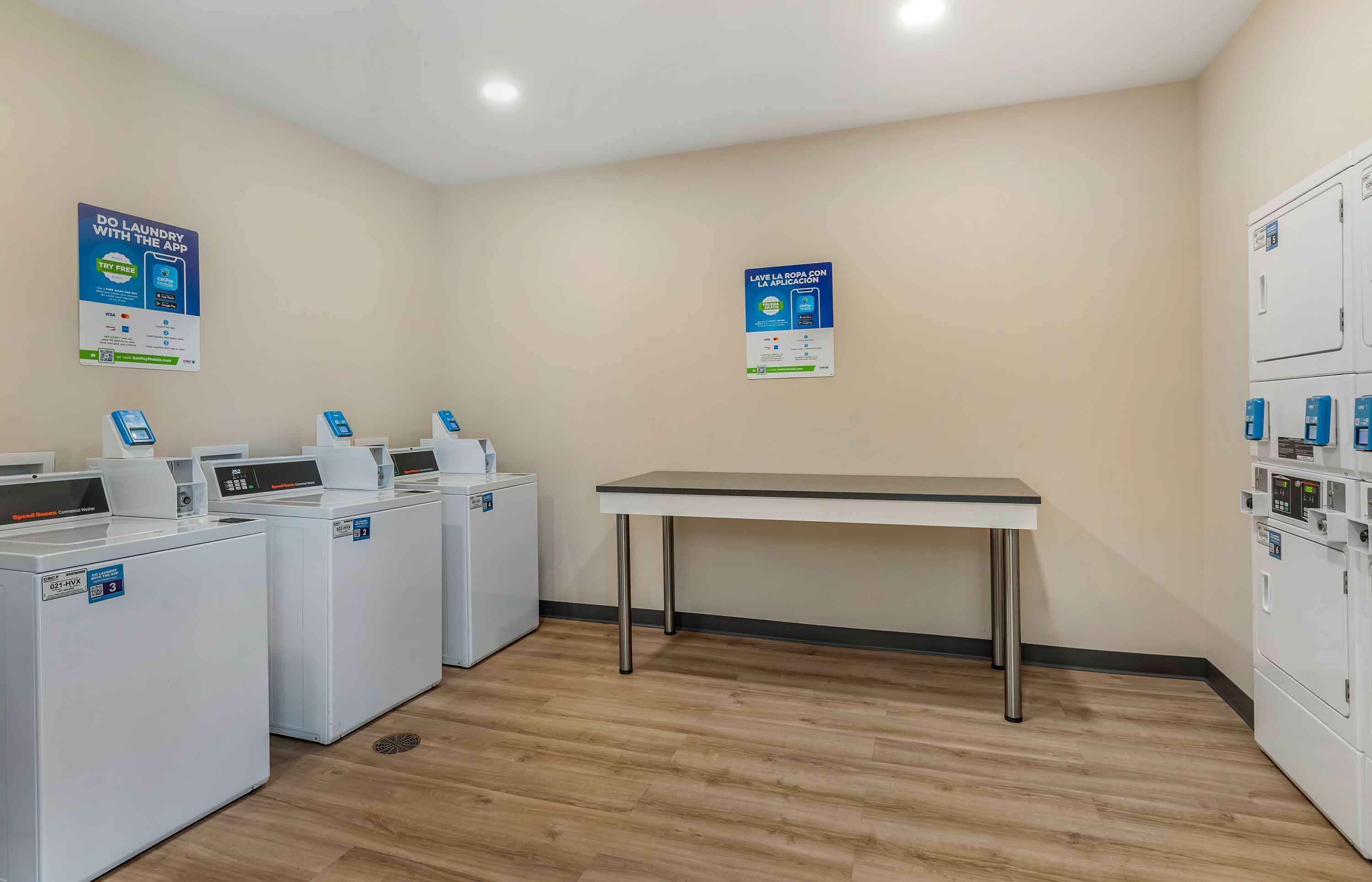 A laundry room with multiple washing machines, a table, and two posters on the walls.