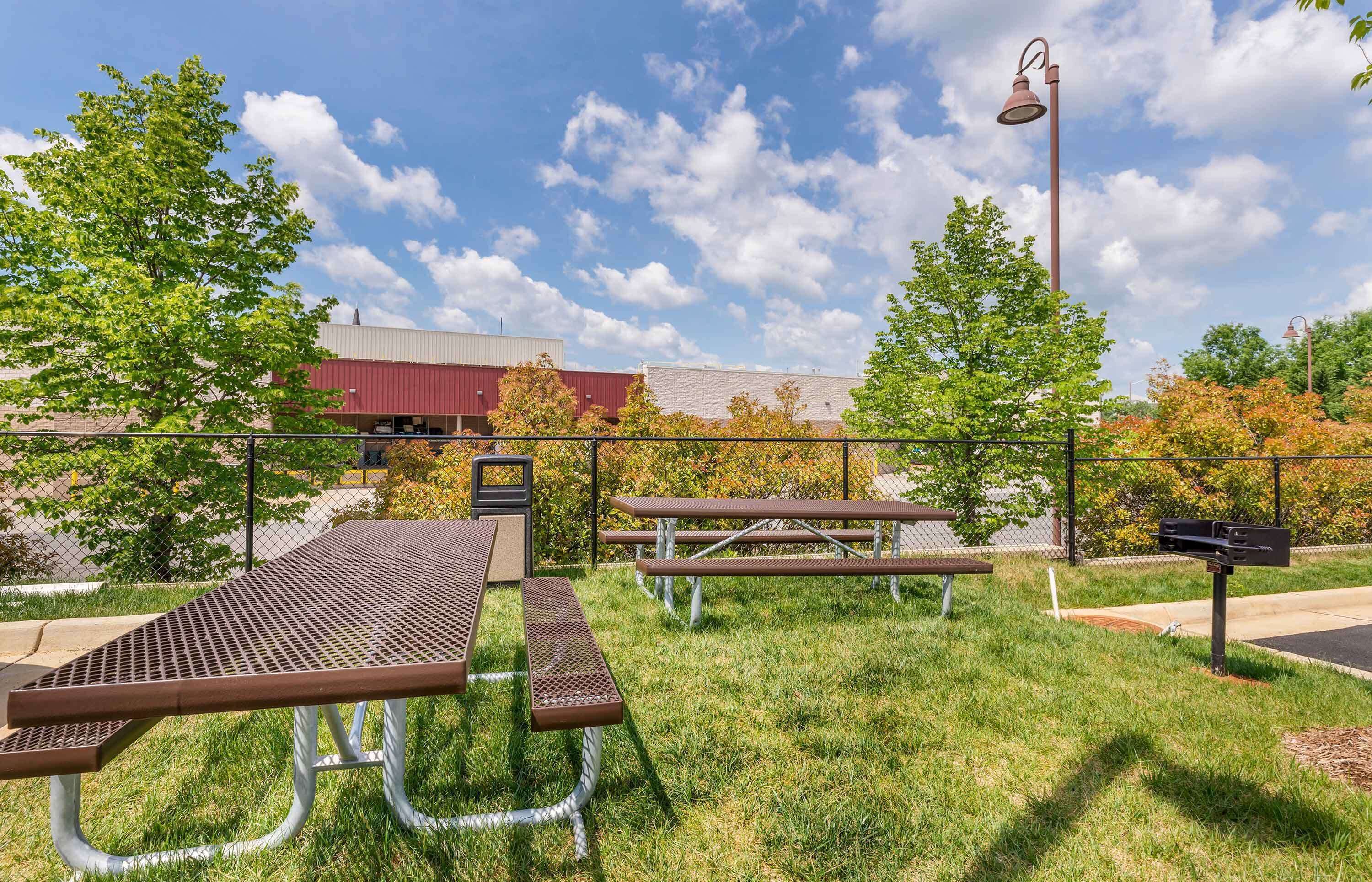 An outdoor picnic area with metal tables and benches, a fence, and a building in the background.