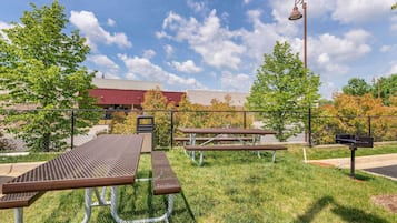 An outdoor picnic area with metal tables and benches, a fence, and a building in the background.
