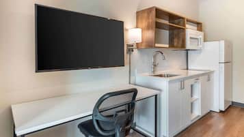 A compact kitchen with a white countertop, a microwave, a refrigerator, and a wooden shelf above the sink.
