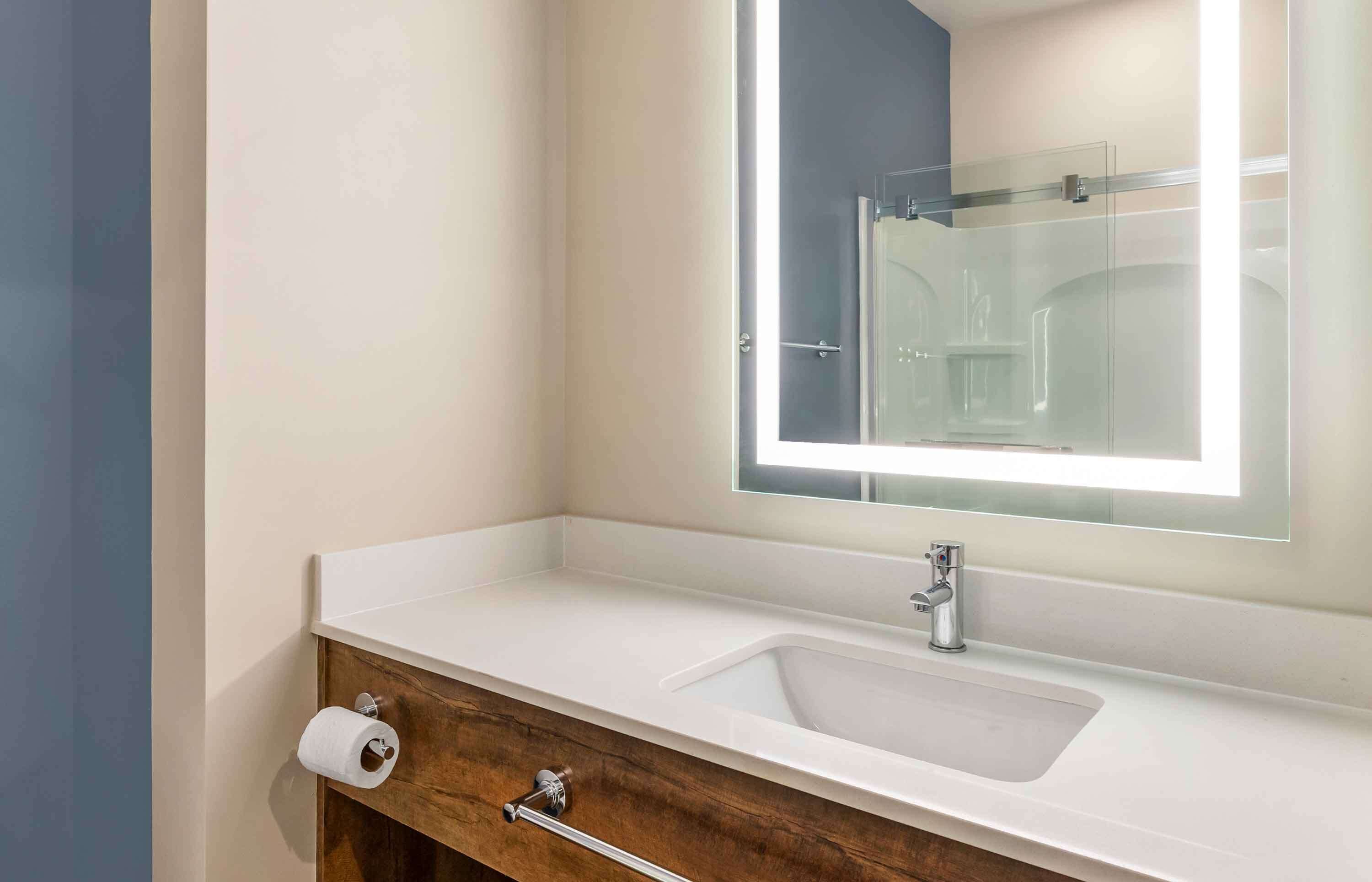 A bathroom with a white sink, a wooden vanity, a mirror with a light, and a toilet paper holder.