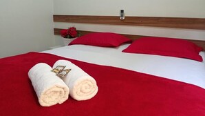 A neatly made bed with red and white bedding, two red pillows, and two towels with a gold emblem. - Pousada Hits Foz (Foz do Iguaçu)