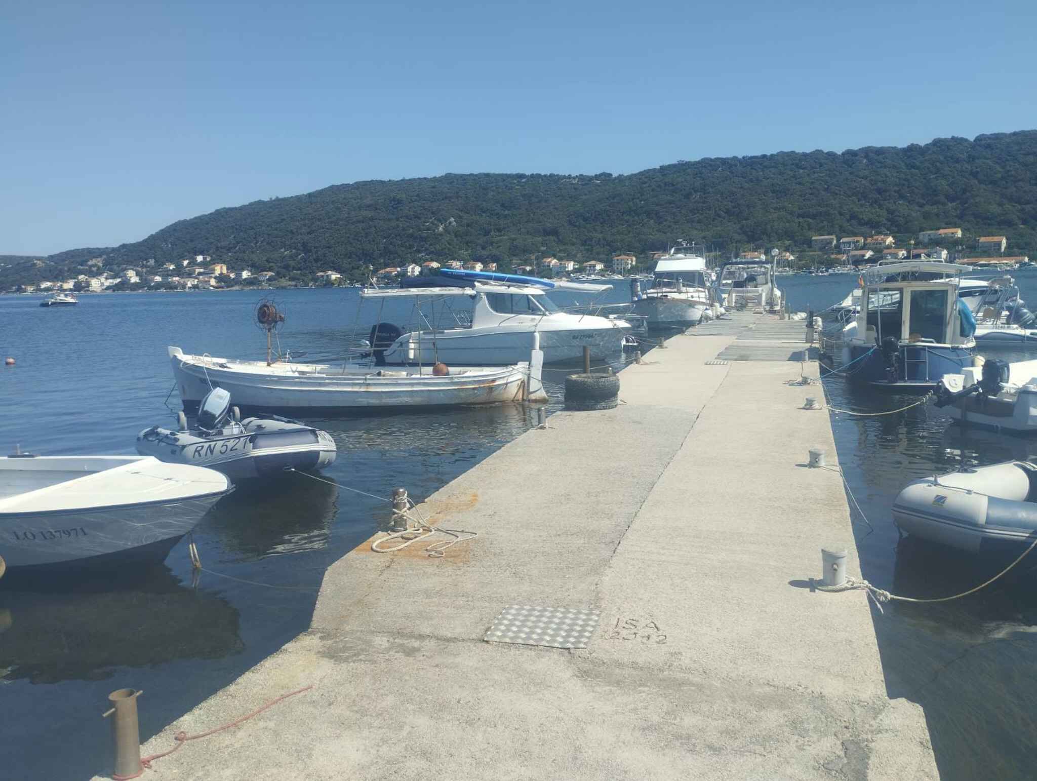 Una marina con barcos amarrados a lo largo de un muelle de hormigón, rodeada por un paisaje montañoso y un pueblo pequeño al fondo.