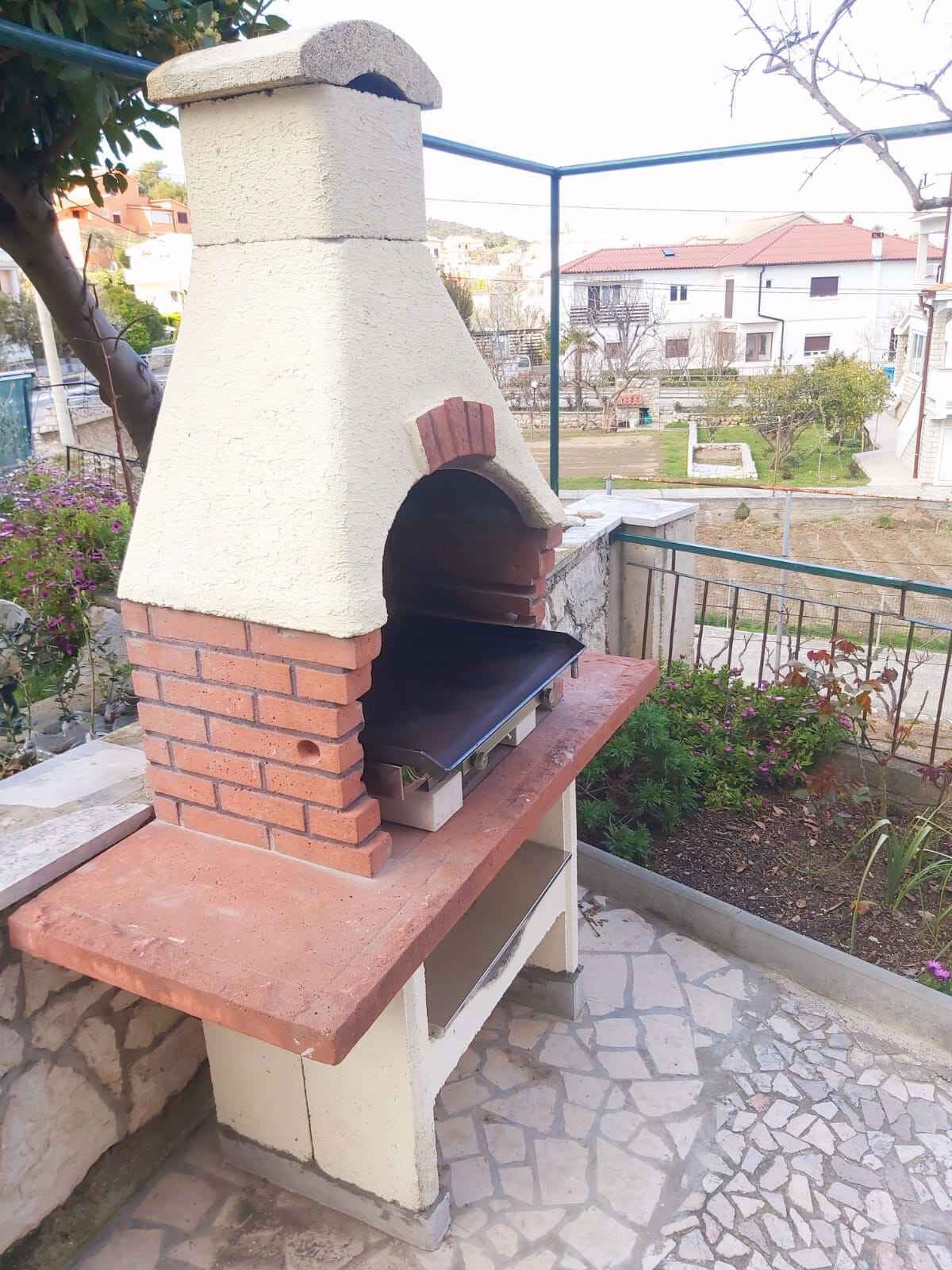 Un four extérieur en briques, doté d’une cheminée, installé sur une terrasse carrelée avec une rambarde en métal, et offrant une vue sur un quartier résidentiel en arrière-plan.