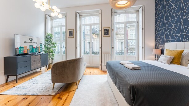 A modern hotel room with a bed, a TV on a dresser, a chair, and a large window. - GuestReady - Amazing Studio in the heart of Porto (Porto)