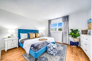 Ein modernes Schlafzimmer mit blauem Kopfteil, einem großen Bett, einem Nachttisch mit Lampe, einem Beistelltisch, einem Fenster mit Vorhängen, einer Topfpflanze und einem dekorativen Korb.