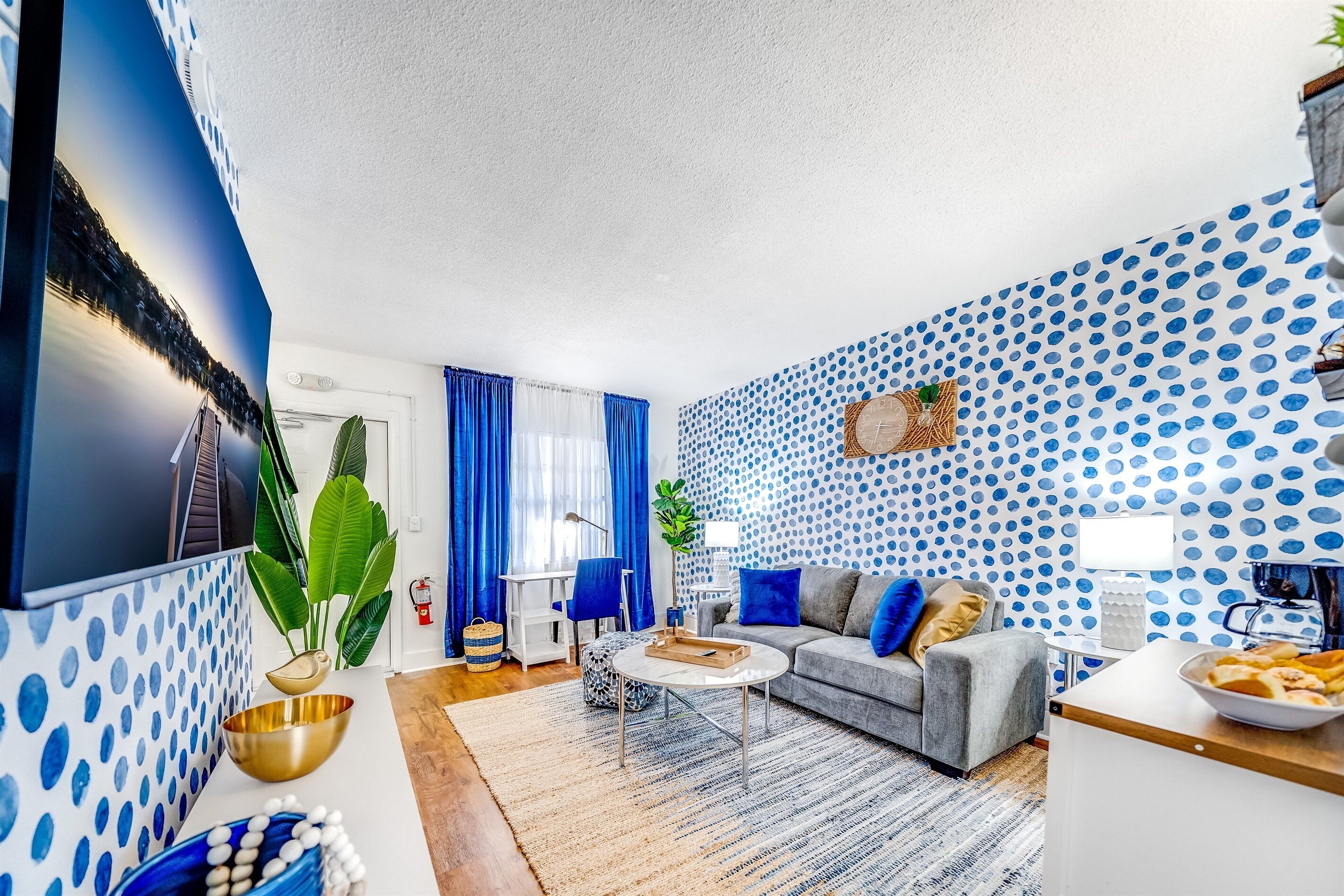 A modern living room with a blue polka dot wallpaper, a grey sofa, a coffee table, and a TV mounted on the wall.
