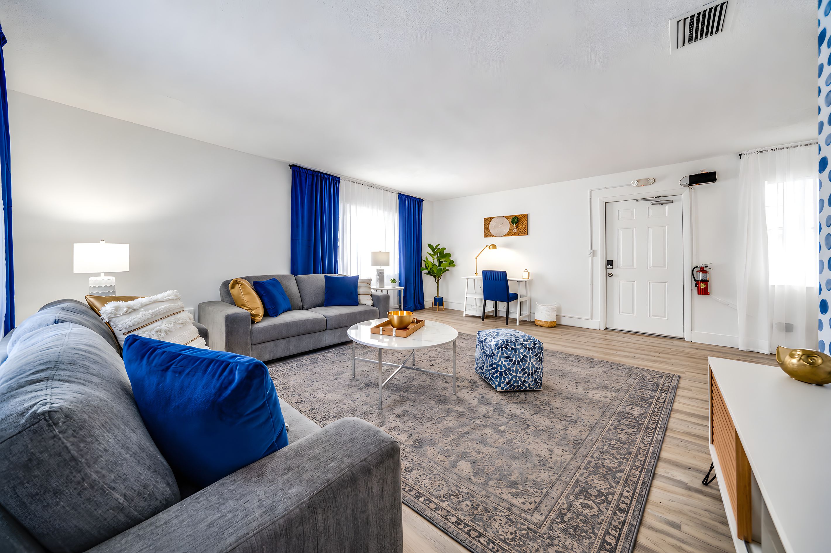 A modern living room with a grey sofa, a coffee table, and a blue patterned ottoman. There is a white door and a window with blue curtains.