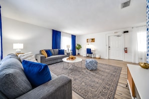 Ett modernt vardagsrum med en grå soffa, ett soffbord och en blå, mönstrad pall. Det finns en vit dörr och ett fönster med blå gardiner.