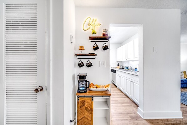 A kitchen with white cabinets, a coffee station, and a wooden table with a coffee maker.