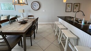 A modern kitchen with a wooden dining table, white bar stools, a large clock, pendant lights, and a tiled floor.