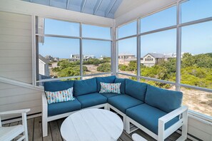 A covered outdoor seating area with a blue sofa and white table, large windows, and a view of houses and greenery.