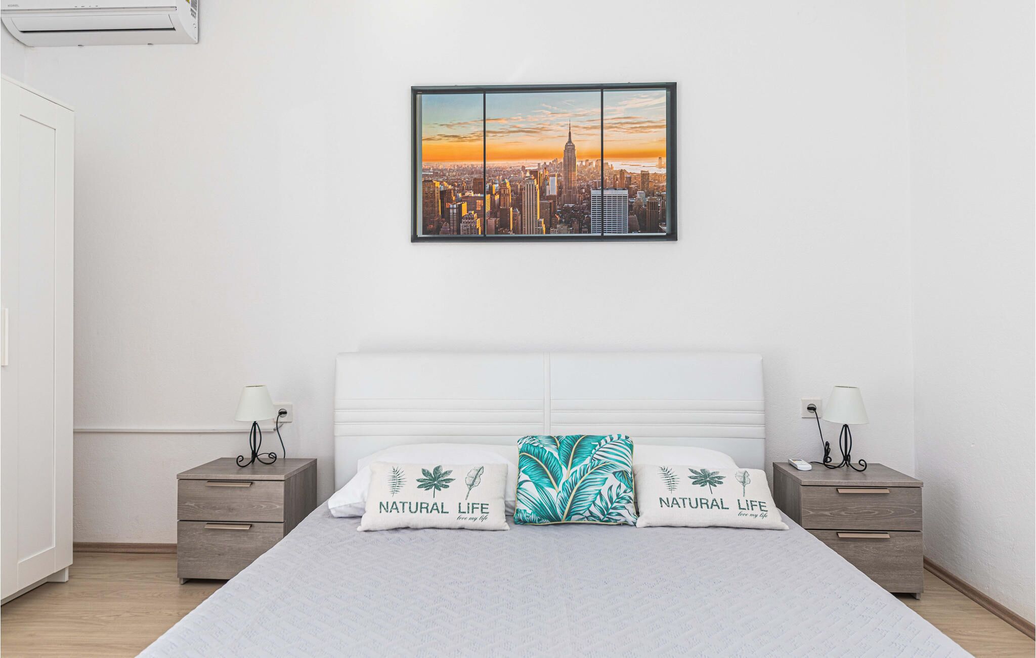 A bedroom with a large bed, two nightstands, a cityscape painting, and a view of a city skyline.