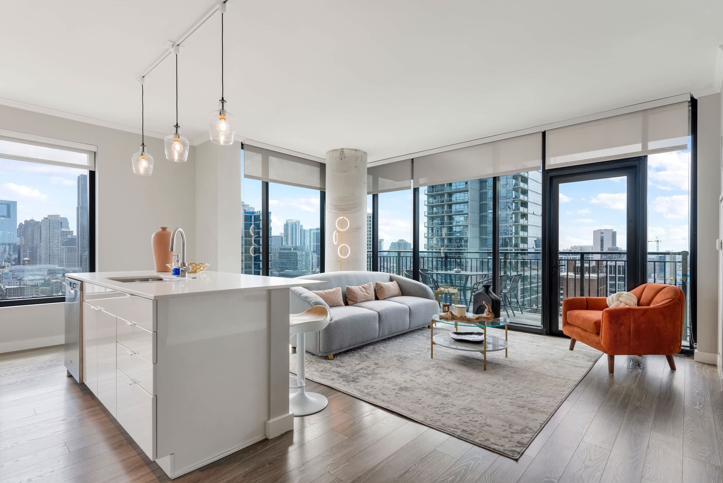 Un apartamento moderno con una isla de cocina, un sofá gris, un sillón naranja y grandes ventanales con vistas a la ciudad.