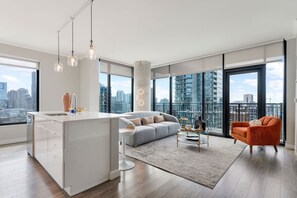 Un apartamento moderno con una isla de cocina, un sofá gris, un sillón naranja y grandes ventanales con vistas a la ciudad.