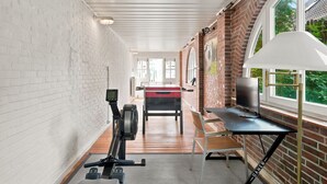 A room with a brick wall, a large window, a desk, a chair, and exercise equipment.