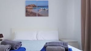 A hotel room with a bed, pillows, a nightstand, a lamp, and a painting of a coastal scene. - Dunasamor near to the Dunes by iRent Fuerteventura (Corralejo)