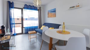 A modern living room with a sofa, a dining table with chairs, a chandelier, and a view through a large window. - Dunasamor near to the Dunes by iRent Fuerteventura (Corralejo)