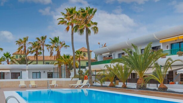 A swimming pool with lounge chairs and palm trees, surrounded by buildings with a Mediterranean architectural style. - Dunasamor near to the Dunes by iRent Fuerteventura (Corralejo)