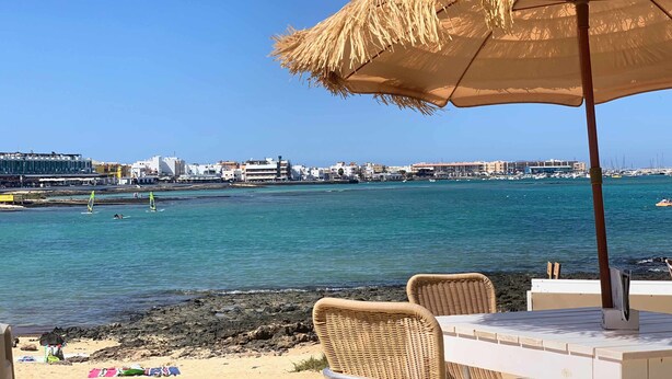 A beachside table with a straw umbrella, wicker chairs, and a view of the sea and cityscape. - Semicorchea Corralejo Center iRent Fuerteventura (Corralejo)
