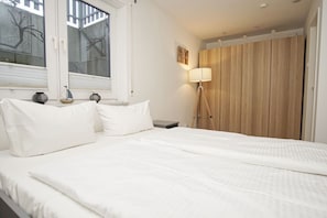Ein ordentlich bezogenes Bett mit weißer Bettwäsche, eine Nachttischlampe und ein Fenster mit Jalousien.