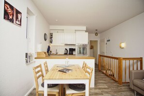 A compact kitchen with white cabinets, a wooden dining table, and a small island with a coffee machine. - Cozy vacation apartment near the harbor - FeWos am Hochzeitsberg FeWo 08 - FeWos (Breege)