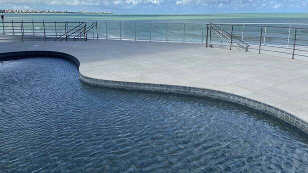 A swimming pool with a curved edge, a tiled deck, and a railing overlooking a body of water. - Bela Hospedagem - Vista piscina e mar (João Pessoa)