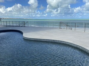 A swimming pool with a curved edge, a tiled deck, and a railing overlooking a body of water. - Bela Hospedagem - Vista piscina e mar (João Pessoa)