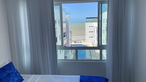A hotel room with a bed, white curtains, and a view of the beach and ocean through a large window. - Bela Hospedagem - Vista piscina e mar (João Pessoa)