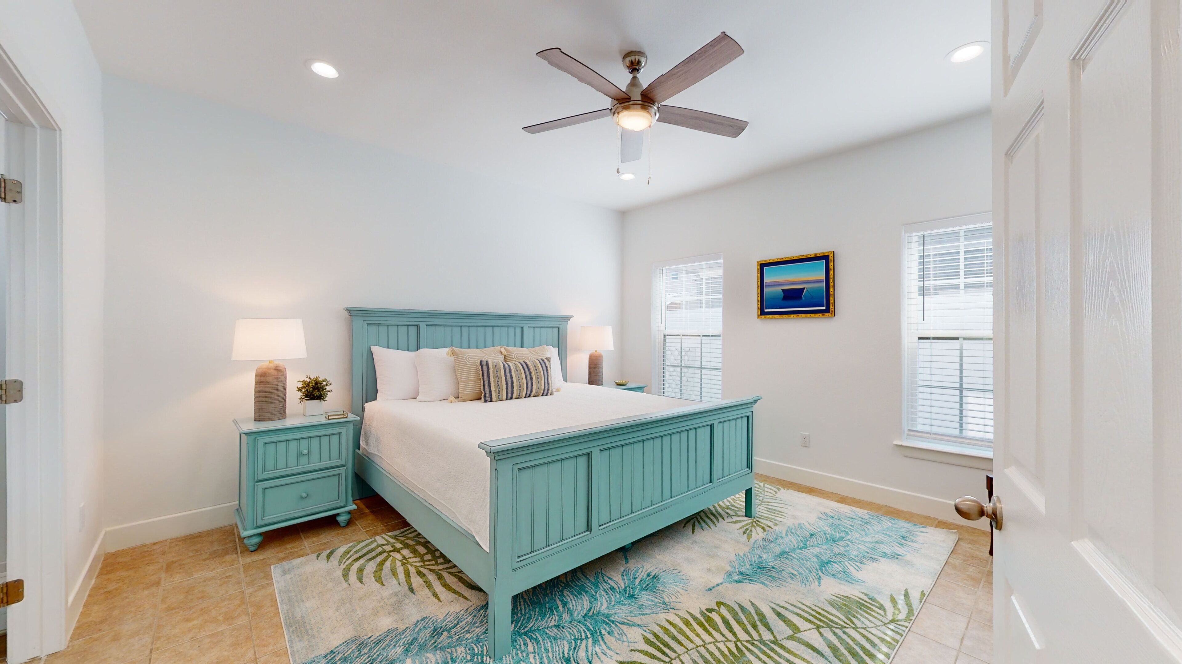 A bedroom with a teal bed, two bedside tables, a ceiling fan, and a window with blinds.
