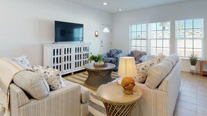 Un salon avec un ventilateur de plafond, un téléviseur, un meuble blanc, un canapé, un fauteuil à motifs et une lampe sur une table d’appoint.